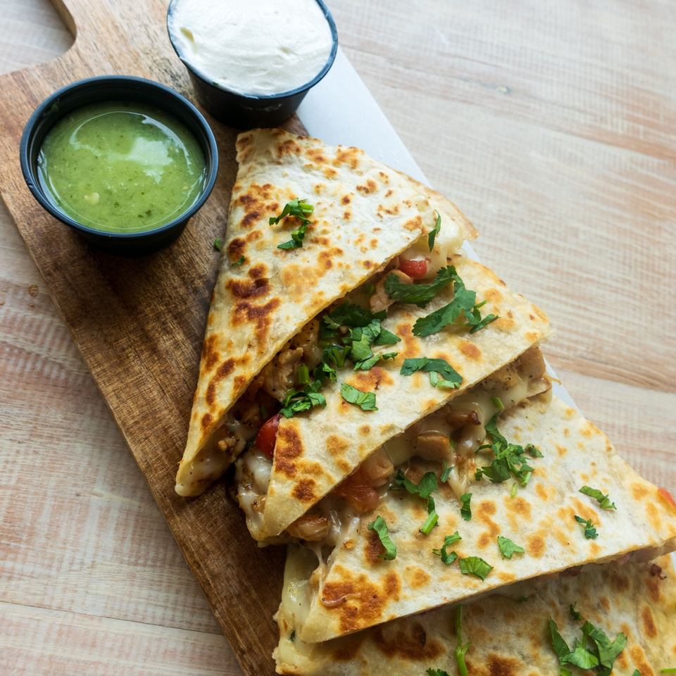 Chicken quesadillas with melted cheese, cilantro, green salsa, and sour cream on a wooden board.