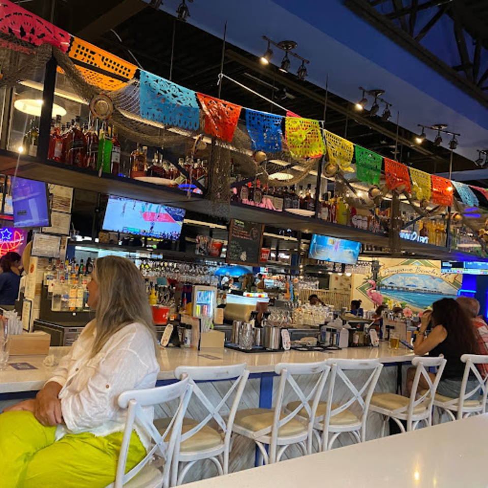 Patrons seated at a lively bar with colorful decorations, liquor bottles, and TVs showing sports.