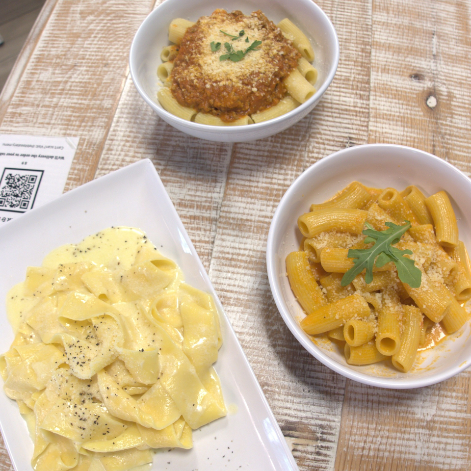 Three pasta dishes on a rustic wooden table, including pappardelle, rigatoni with bolognese, and rigatoni with cheese sauce.