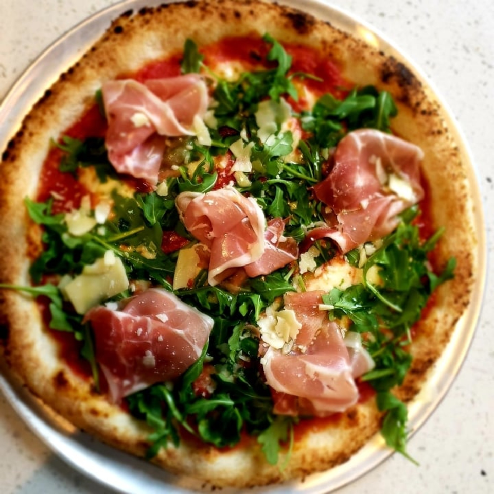 A delicious pizza from The Bite Eatery, topped with prosciutto, fresh arugula, and shaved parmesan.