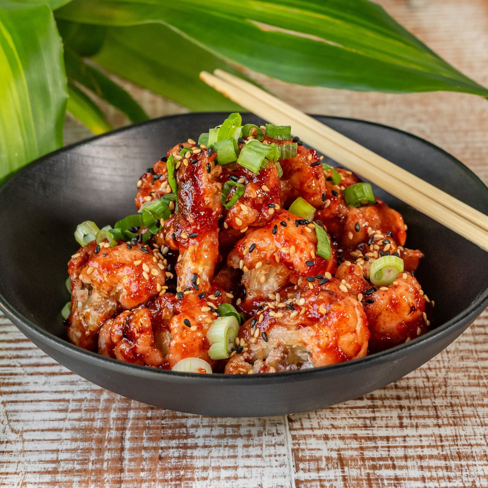 Sweet and sour chicken bites with sesame seeds and green onions in a black bowl with chopsticks.
