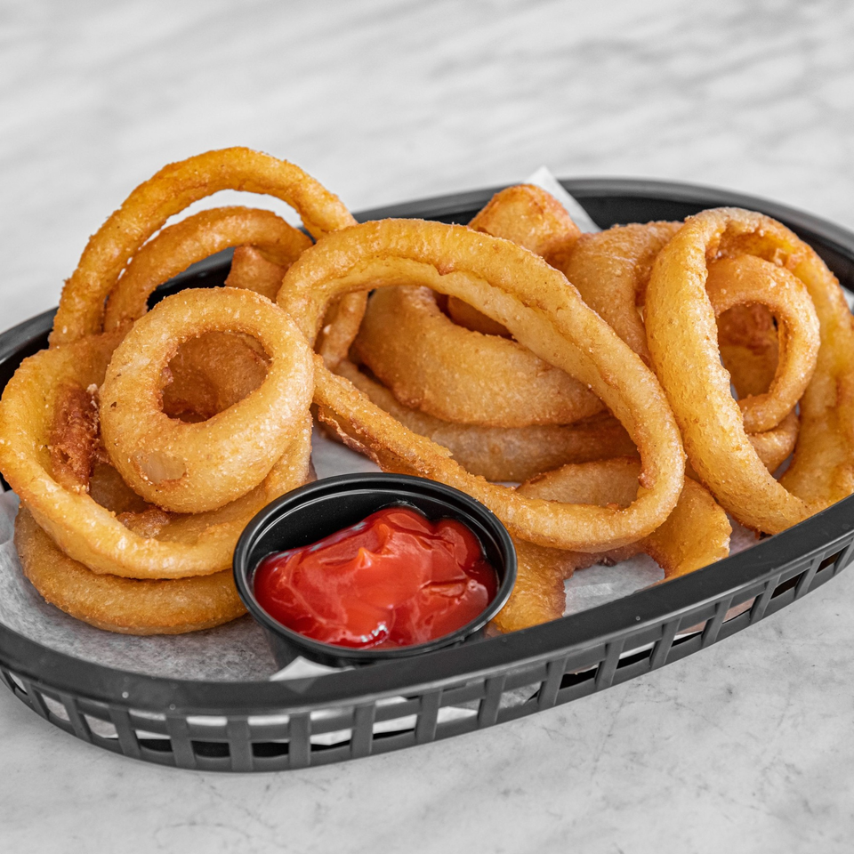 A black basket filled with golden-brown onion rings and a small cup of red ketchup.